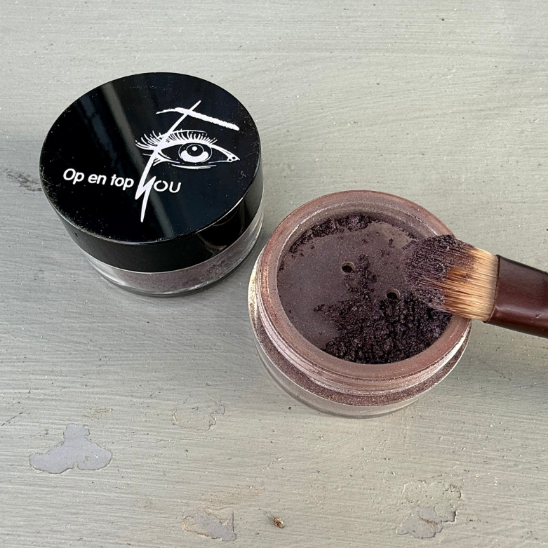 Een potje met minerale oogschaduw in een glanzende aubergine/amora tint. De losse poedertextuur heeft een subtiele glans en een diepe bruin-paarse kleur. Het potje is open, waardoor de rijke pigmentatie en fijne, poederachtige structuur goed zichtbaar zijn. De kleur straalt luxe en warmte uit, ideaal voor een elegante ooglook.
